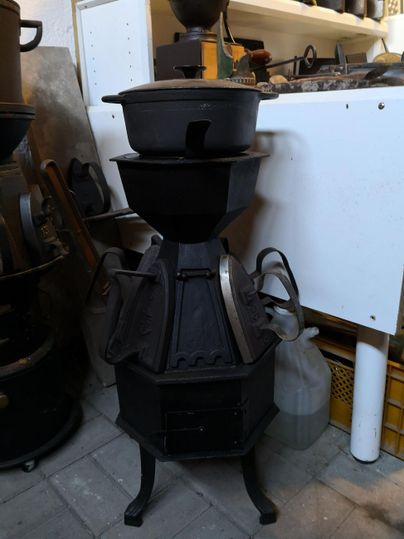 Ein rustikaler, schwarzer Gusseisenofen steht in einer Werkstatt. Der Ofen hat mehrere Schichten und einen stabilen Fuß.