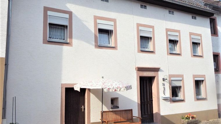 A simple two-story house with white walls and a red frame. In front of the house is a bench and a sun umbrella.
