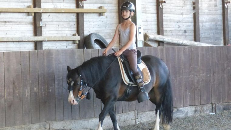 Een kind met een helm rijdt op een pony in een rijhal. De omgeving is van hout en heeft een rustieke uitstraling.