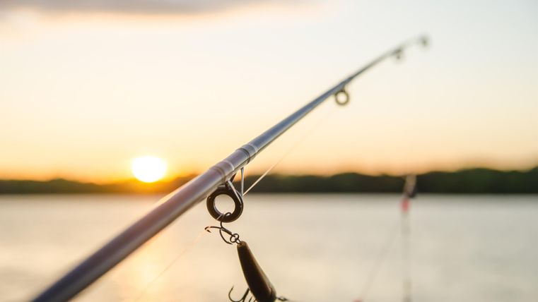 A fishing rod stands in the foreground while the sun sets on the horizon. The calm water horizon and the gentle colors of the sunset create a relaxed atmosphere.
