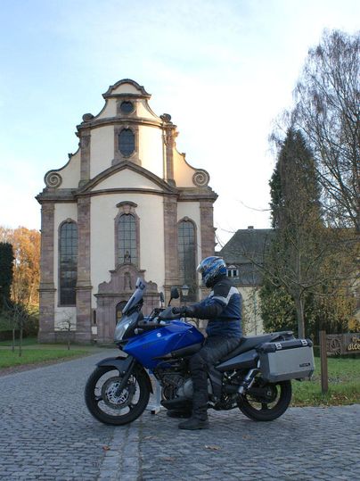 Ein Motorradfahrer sitzt auf einem blauen Motorrad vor einer beeindruckenden Kirche. Die Umgebung ist grün und die Architektur der Kirche ist historisch.