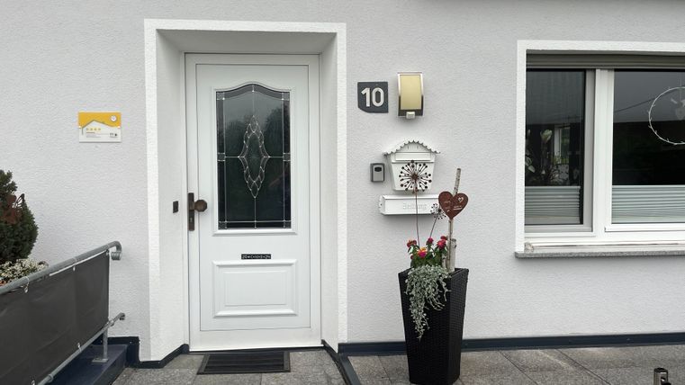 Eine freundliche Eingangstür mit einer schönen Veranda und modernen Fenstern. Neben der Tür steht ein Blumenkübel mit dekorativen Pflanzen.