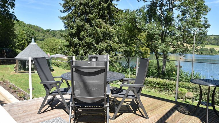 Ein schöner Außenbereich mit einem Tisch und Stühlen auf einer Terrasse. Im Hintergrund sieht man einen See und viel grüne Natur.