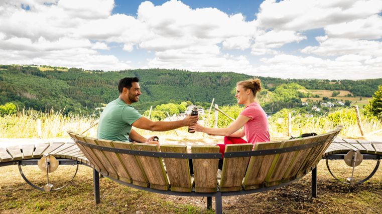 Zwei Personen sitzen auf einer Holzbank mit Blick auf eine grüne Landschaft und stoßen mit Thermosflaschen an.