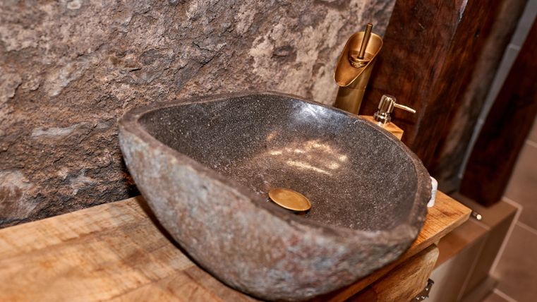 An elegant stone sink is placed on a wooden shelf. In the background, natural stones are visible, creating a rustic atmosphere.