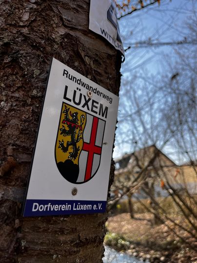 A sign with the coat of arms of Lüxem shows the circular hiking trail. In the background, there are trees and a clear sky.