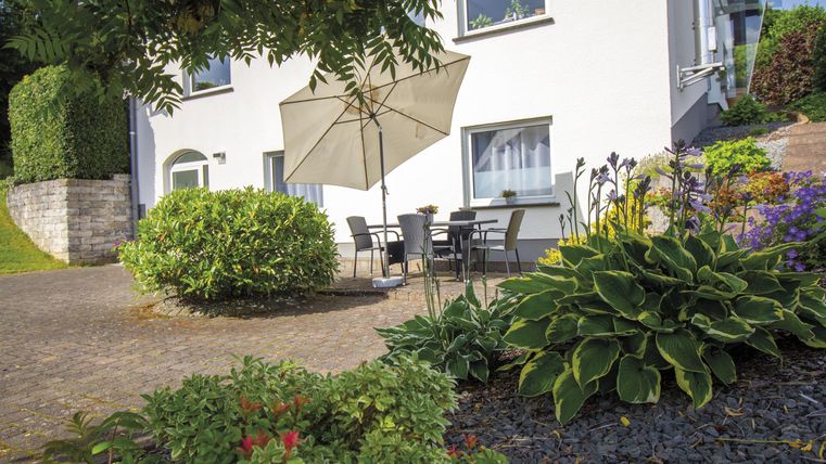 A cozy outdoor area with a table, chairs, and a sun umbrella. Surrounded by colorful plants and bushes.