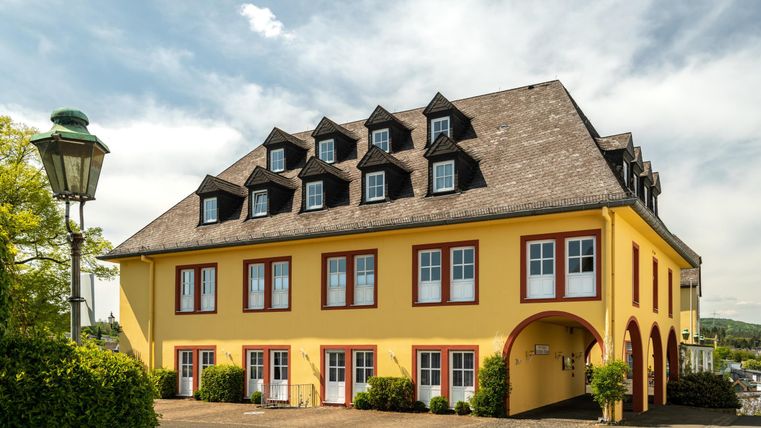 Ein gelbes Gebäude mit vielen Fenstern und einem Satteldach. Vor dem Haus sind dekorative Pflanzen und eine Straßenlaterne zu sehen.