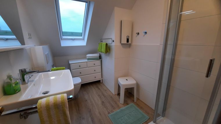 A modern bathroom with a shower and a sink. There are bright walls, wooden flooring, and fresh towels in green colors.