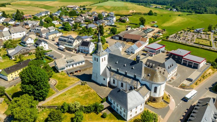 A picturesque village with a church in the center, surrounded by green hills. The architecture is typical of the rural setting.