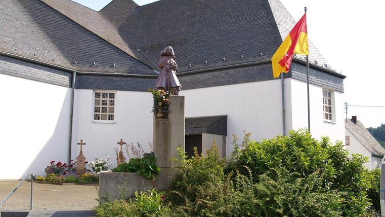 Eine Kirche mit einem Denkmal und einer Flagge im Vordergrund. Die Umgebung ist grün und gepflegt.
