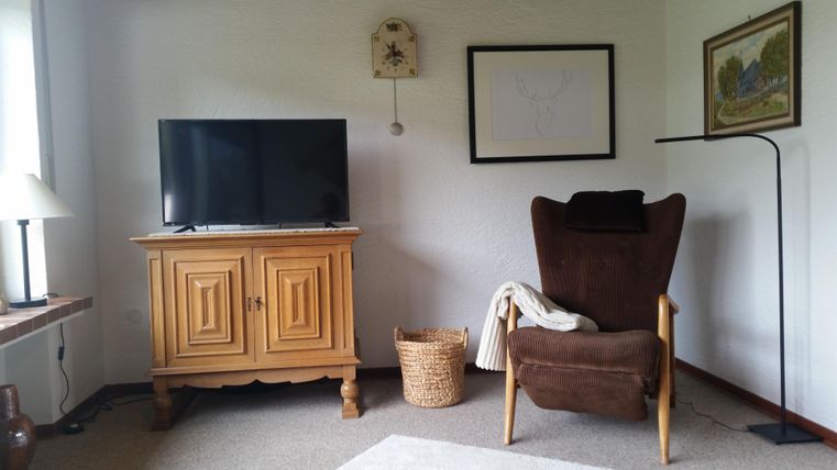 Ein gemütliches Wohnzimmer mit einem Fernseher, einem Holzmöbel und einem bequemen Sessel. Im Raum ist auch ein Korb und ein Stehlampe sichtbar.