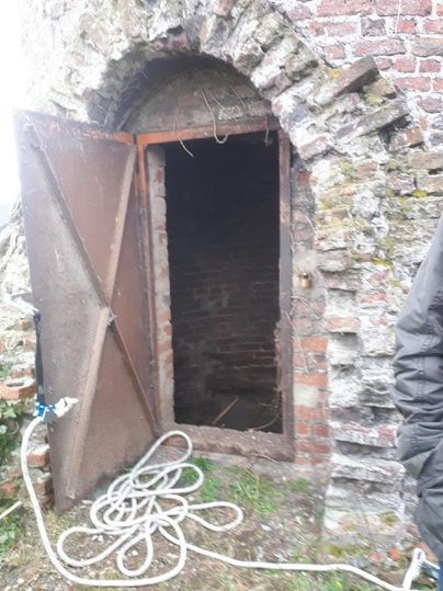 Een oude, open metalen deur leidt naar een donkere kamer. Voor de deur liggen een paar slangen op de grond.