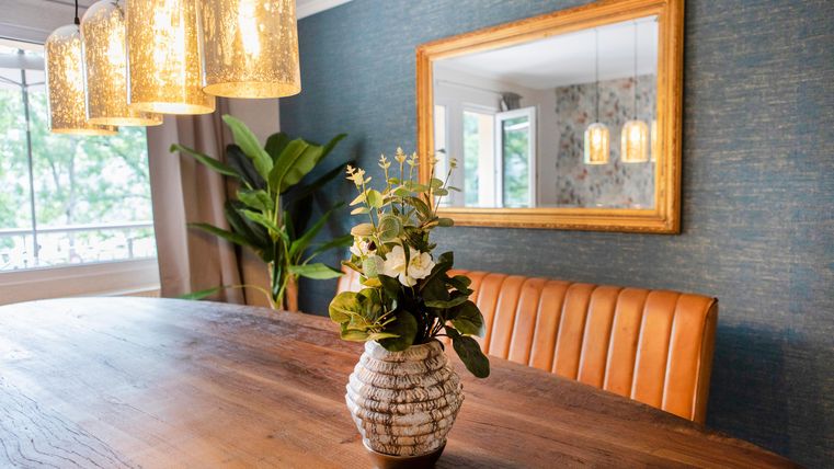 Ein eleganter Essbereich mit einem holzigen Tisch und einer schönen Blumenvase. An der Wand hängt ein großer Spiegel und es gibt moderne Hängeleuchten.