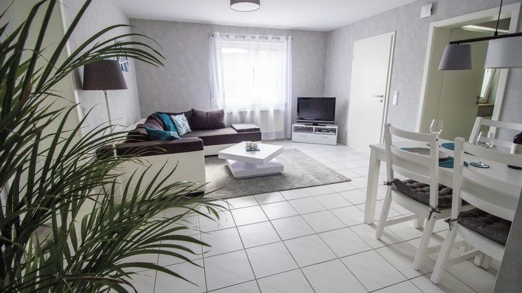 A modern living room with light walls and a large window. There is a cozy couch, a television, and a dining table with chairs.