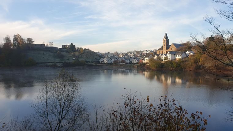 Een rustig landschap met een meer en uitzicht op een pittoresk dorp. Op de achtergrond is een kerktoren en een beboste heuvelrug te zien.