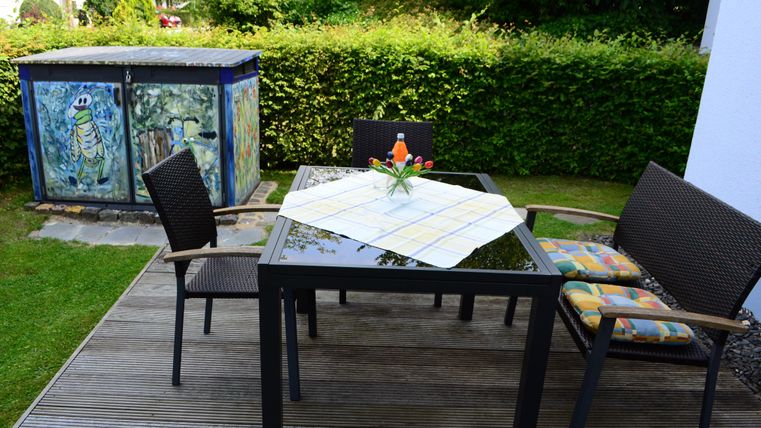 A cozy garden table with chairs and a tablecloth. In the background, there is a green hedge and a brightly painted shed.