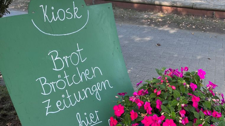 Een kiosk bord met de tekst "Brood, broodjes, kranten". Ernaast bloeien roze bloemen.