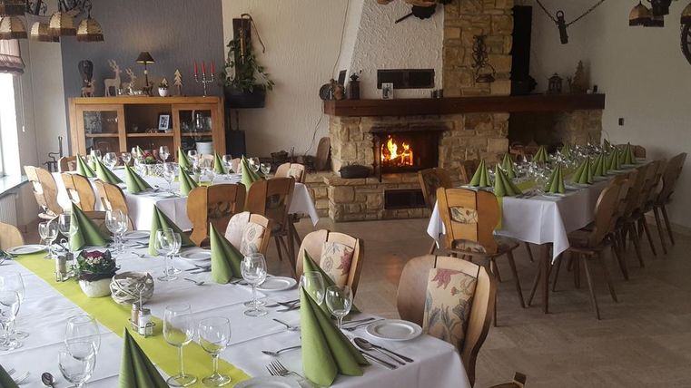 Ein gemütlicher Speisesaal mit festlich gedeckten Tischen und grünen Servietten. Im Hintergrund strahlt ein offenes Feuer im Kamin angenehme Wärme aus.