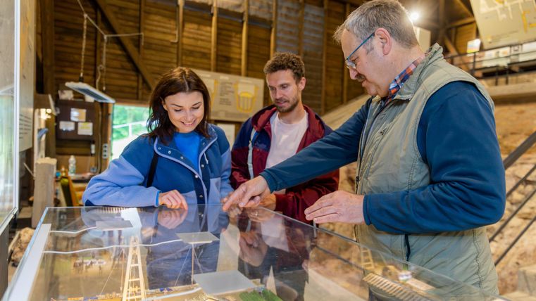 Drei Personen betrachten ein Modell in einem Ausstellungsraum. Zwei junge Erwachsene hören aufmerksam zu, während ein älterer Mann erklärt.