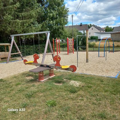 Een speeltuin met een wip, schommel, klimwand en bogen op grind, omgeven door bomen en huizen.