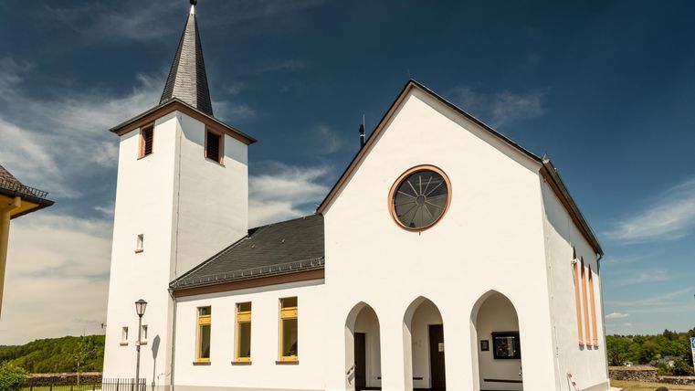 Eine weiße Kirche mit einem spitzen Turm und großen Fenstern. Der Himmel ist klar und blau, umgeben von grüner Landschaft.