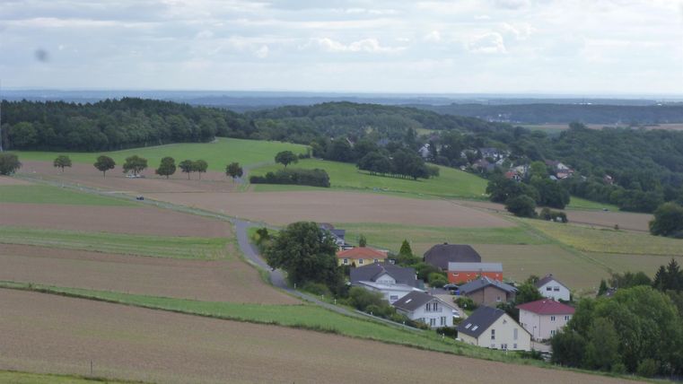 Eine ländliche Landschaft mit sanften Hügeln und Feldern. Im Vordergrund ist ein kleines Dorf mit mehreren Häusern zu sehen.