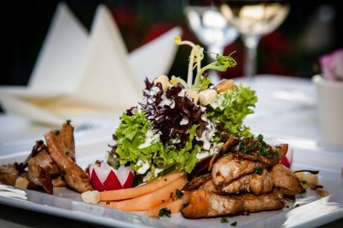Ein kunstvoll angerichtetes Gericht mit Fleisch, Salat, Radieschen und Melone auf einem weißen Teller, im Hintergrund ein Glas Weißwein.
