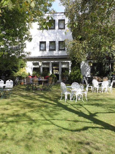 Ein schönes Gartengelände mit einer weißen Fassade im Hintergrund. Auf der Wiese stehen mehrere Gartenstühle und -tische unter schattenspendenden Bäumen.