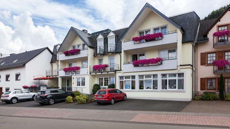 Ein mehrstöckiges Wohngebäude mit Balkonen und blühenden Pflanzen. Vor dem Gebäude stehen mehrere Autos und der Himmel ist teilweise bewölkt.
