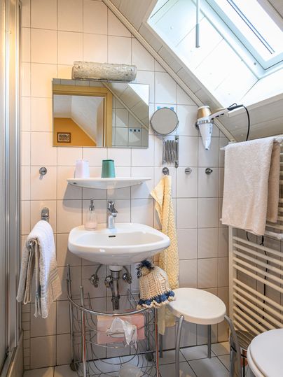 Ein kleines Badezimmer mit einer Dusche, einem Waschbecken und einem Spiegel. Es gibt helle Fliesen und Handtücher an der Wand.