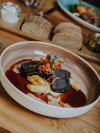 Ein eleganter Teller mit gebratener Blutwurst, serviert auf Püree und einer Soße. Das Gericht ist mit knusprigen Zwiebeln und frischen Kräutern garniert.
