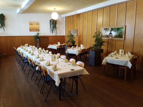 A festively decorated hall with long tables, white tablecloths, flowers and plants on the walls. Wooden walls and parquet flooring create a warm atmosphere.