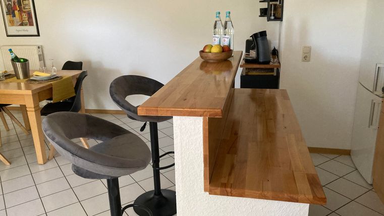 A modern kitchen with a wooden bar and high chairs. There are bottles of water and a fruit basket on it.