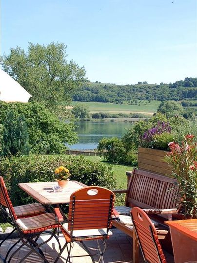 Ein gemütlicher Außenbereich mit Tischen und Stühlen bietet einen Blick auf einen ruhigen See und grüne Hügel im Hintergrund. Die Atmosphäre ist entspannt und einladend.