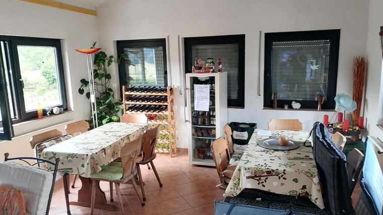 Ein heller Essbereich mit mehreren Tischen und Stühlen. Die Wände sind mit Fenstern ausgestattet und eine Weinregal ergänzt die Einrichtung.