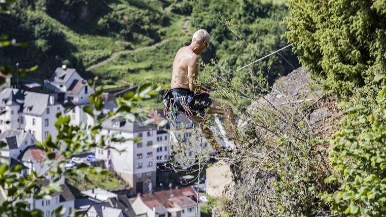 Ein Mann klettert an einem Felsen, gesichert mit Seilen. Im Hintergrund ist ein Dorf mit weißen Häusern und grüner Landschaft zu sehen.