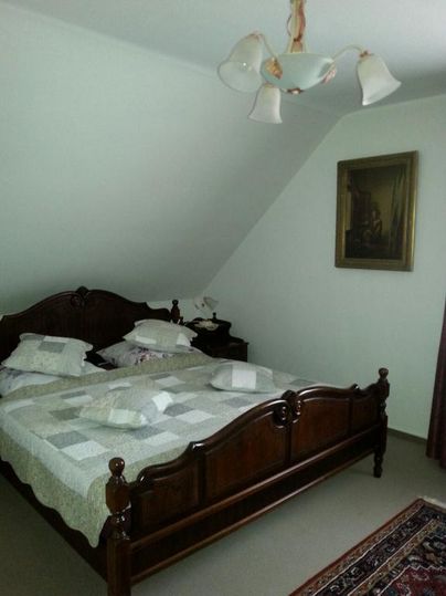 Ein gemütliches Schlafzimmer mit einem großen, hölzernen Bett und mehreren Kissen. An der Wand hängt ein Gemälde, und ein Teppich liegt auf dem Boden.