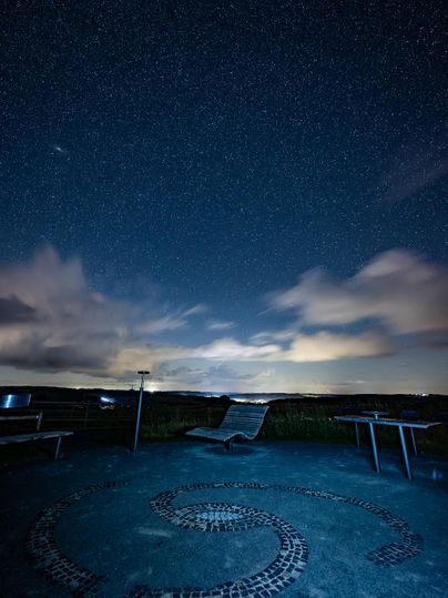 Ein klarer Nachthimmel voller Sterne über einem ruhigen Beobachtungsbereich. Im Vordergrund sind Sitzmöglichkeiten und ein kunstvolles Muster auf dem Boden zu sehen.