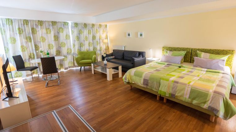 Ein helles Zimmer mit einem Doppelbett, einer Sitzecke und einem Tisch. Die Wände sind mit bunten Vorhängen und einem warmen Holzboden dekoriert.