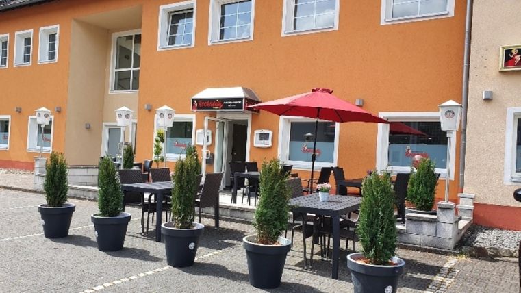 Een modern gebouw met oranje gevel en grote ramen. Buiten staan tafels met stoelen en een rode parasol, omringd door planten in zwart-witte potten.