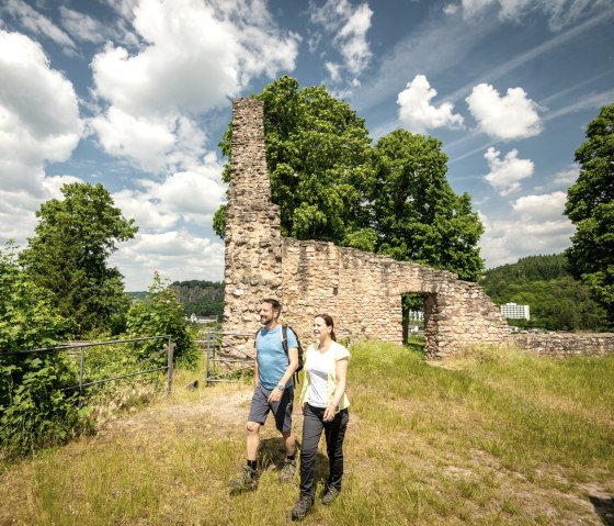 Zwei Wanderer spazieren an einer alten Steinmauer vorbei, umgeben von grünen Bäumen und einem blauen Himmel mit Wolken., © Eifel Tourismus GmbH, Dominik Ketz
