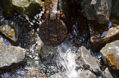 Ein Wasserlauf fließt über Steine. In der Mitte ist eine runde, rostige Abdeckung sichtbar.