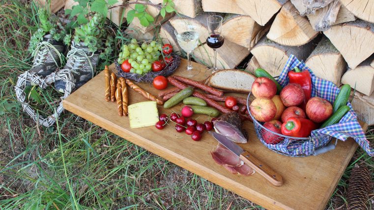 A rustic table with an assortment of fresh fruits, vegetables, cheese, and cold cuts. In the background, there are firewood and a glass of wine.