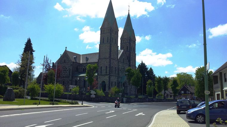 Een indrukwekkende kerk met twee hoge torens tegen een blauwe lucht. De omgeving is groen en uitnodigend, met een straat op de voorgrond.