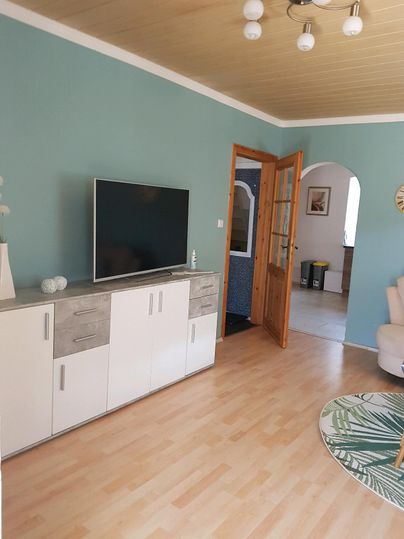 Een lichte woonkamer met een blauwe muur en een moderne tv-kast. Op de achtergrond is er een open deur die naar een aangrenzende ruimte leidt.