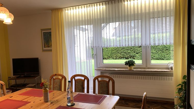 Ein gemütlicher Essbereich mit einem Holztisch und Stühlen. Große Fenster mit hellen Vorhängen bieten Ausblick auf den Garten.