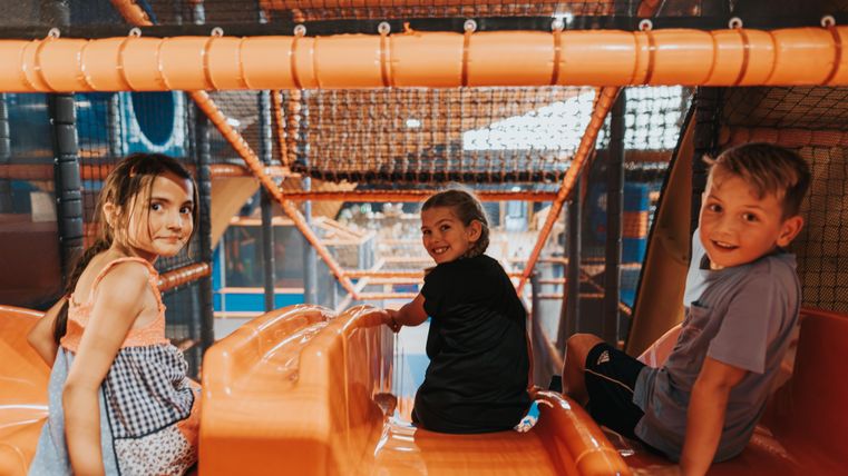 Drei Kinder spielen in einem bunten Spielplatz mit Rutschen. Sie haben Spaß und lächeln in die Kamera.