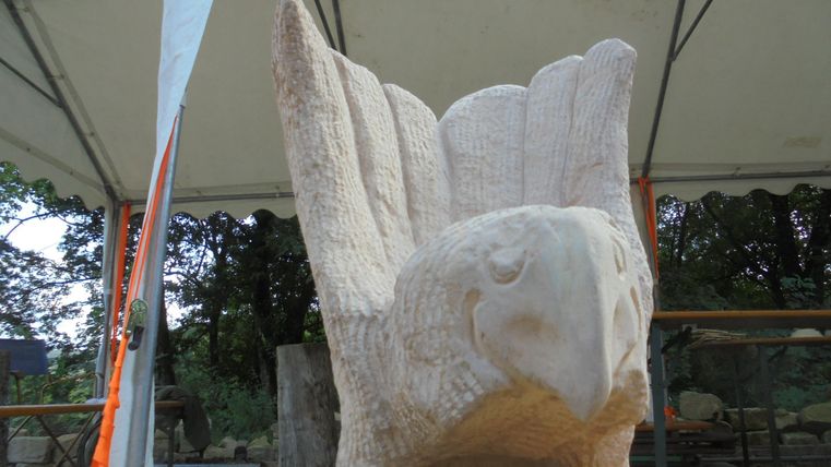 An artistic sculpture of a bird with prominent wings made of stone. The statue stands on a table and is protected by a tent canopy.