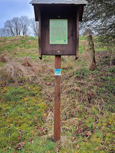 Een informatiebord van hout staat op een grasveld. Op de achtergrond zijn bomen en een bewolkte lucht te zien.